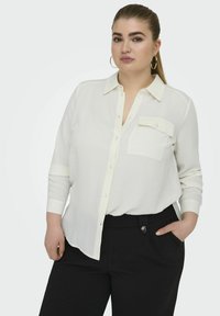 Cămașă albă cu mâneci lungi, cu guler, dintr-un material texturat, și cu un buzunar pe piept, asociată cu pantaloni negri cu talie înaltă, având un detaliu cu un nasture.