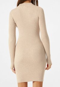 Robe en tricot côtelé beige clair, présentant une silhouette ajustée, des manches longues et un col montant. Texture lisse et design arrive aux genoux.