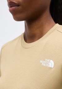 Κοντινό πλάνο ατόμου που φοράει μπεζ υφασμάτινη μπλούζα με λευκό λογότυπο The North Face στο στήθος.