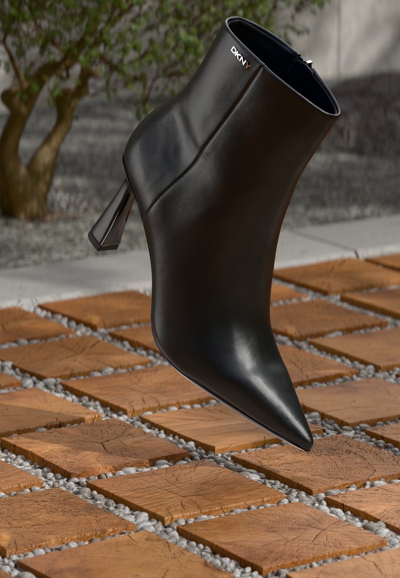 Bota de tornozelo em couro preto com bico pontiagudo e um salto geométrico. Apresenta uma marcação subtil e uma textura suave. O fundo tem azulejos texturizados.