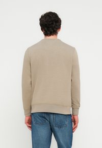 Μπεζ φούτερ με στρογγυλή λαιμόκοψη, ριμπ μανσέτες και τελείωμα, και διακριτική ραφή σε σχήμα V στο πίσω μέρος. Φοριέται με μπλε τζιν παντελόνια.