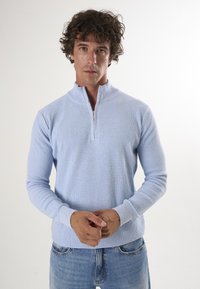 Pull bleu clair en tricot côtelé avec col montant zippé et poignets ajustés, assorti à un jean en denim bleu. Design minimaliste et texture douce.
