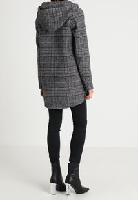 Persona vistiendo un abrigo con capucha de cuadros grises, pantalones negros ajustados y botines negros con tacones de bloque plateados, de pie de espaldas.