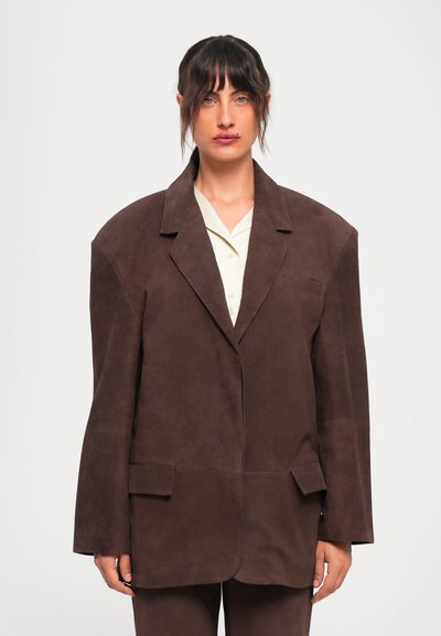 Vrouw met donker haar draagt een oversized bruine suède blazer over een lichtbeige blouse, staande tegen een effen witte achtergrond.