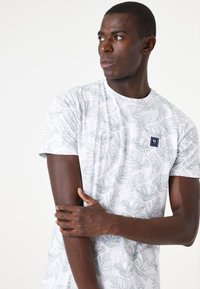 Witte t-shirt met een grijs bladpatroon, ronde halslijn en een klein logo op de borst. Korte mouwen en een ontspannen pasvorm.