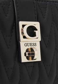 Sac à main en cuir noir matelassé avec un fermoir carré doré portant un logo "G" et le mot "GUESS" embossé sous le détail du verrou.