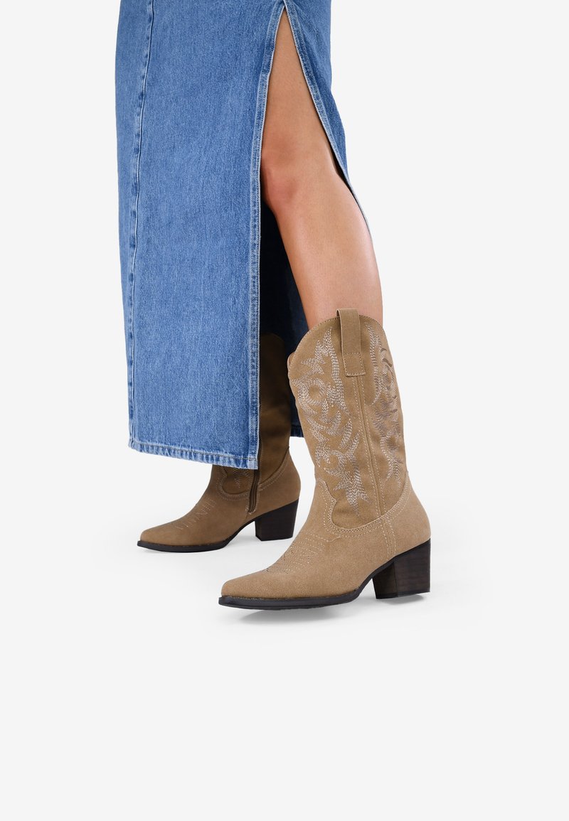 Stivali da cowboy in suede tan con punta appuntita e tacco impilato. Motivi floreali ricamati sulla gamba. Indossati con una gonna di jeans con spacchi laterali.