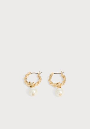 Boucles d'oreilles créoles torsadées en or, chacune avec un grand pendentif en perle blanche sur un fond gris clair.