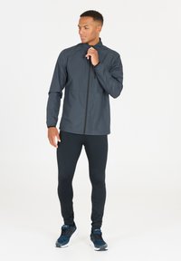 Lättviktsjacka i mörkgrått syntetmaterial, med dragkedja framtill, hög krage och elastiska ärmmuddar, tillsammans med svarta leggings och sneakers.