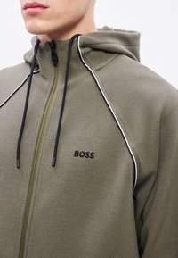 Kurtka na zamek w kolorze oliwkowej zieleni z kapturem, teksturowaną tkaniną, czarnymi sznureczkami i białym przeszyciem, z logo „BOSS” na piersi.