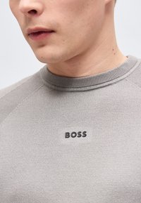 Gros plan sur une personne portant un pull en maille gris clair avec un col rond et le logo "BOSS" sur la poitrine.