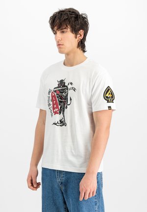 Junger Mann mit dunklem, gewelltem Haar, der ein weißes T-Shirt mit schwarzer Teufelsgrafik und rotem Schild trägt, kombiniert mit blauen Jeans, seitlich stehend.