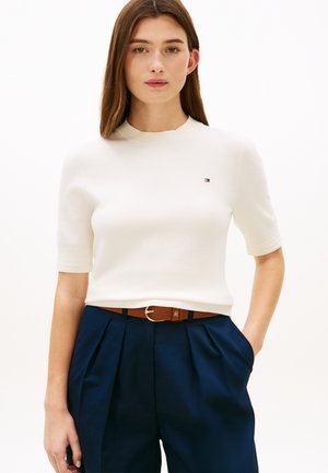 Donna con una maglietta bianca a maniche corte con un piccolo logo, pantaloni plissettati blu navy e cintura marrone, in piedi con una mano in tasca.