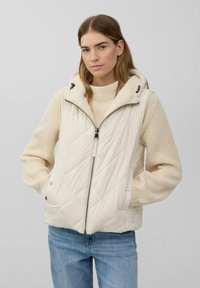 Frau trägt eine cremefarbene gesteppte Weste mit Kapuze über einem beigen Strickpullover und blauen Jeans, steht mit Händen in den Westentaschen.