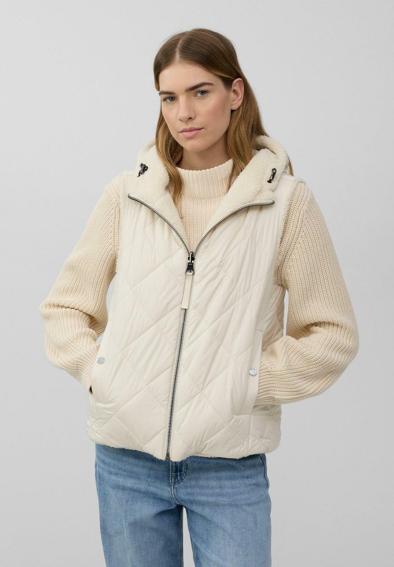 Frau trägt eine cremefarbene gesteppte Weste mit Kapuze über einem beigen Strickpullover und blauen Jeans, steht mit Händen in den Westentaschen.