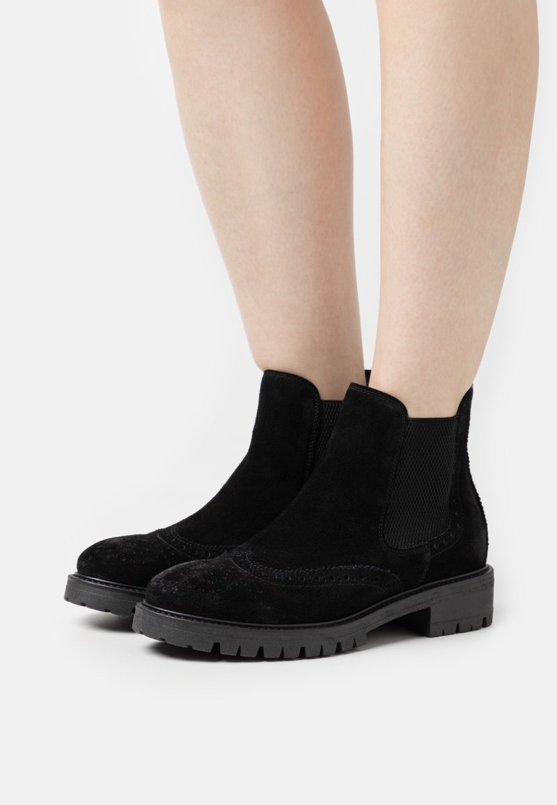 Jambes chaussées de bottines Chelsea en daim noir avec des détails brogue et des semelles épaisses en caoutchouc, se tenant devant un fond blanc.