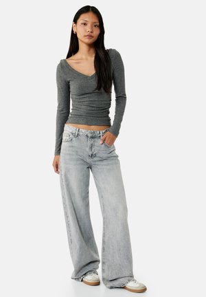 Young woman wearing a fitted grey long-sleeve top, light grey wide-leg jeans, and white sneakers, standing with one hand in pocket.