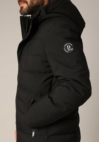 Man in een zwart gewatteerd jack met capuchon, voorzien van een ritszak op de borst en een cirkelvormige "Deeluxe Original" logo patch op de mouw.