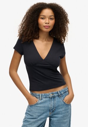 Jeune femme aux cheveux bouclés portant un haut noir à manches courtes et col en V ainsi qu'un jean bleu clair, les mains dans les poches, se tenant devant un fond blanc.