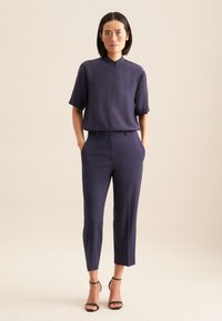 Blusa blu navy con colletto alto, maniche corte e chiusura a bottoni, abbinata a pantaloni blu navy sartoriali e scarpe con cinturino alla caviglia nere.