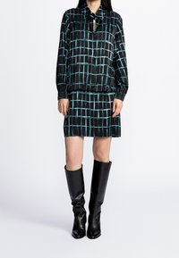 Blusa nera e teal con motivo, colletto e dettaglio a chiave, abbinata a una gonna coordinata. Indossata con stivali in pelle nera al ginocchio.