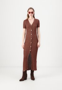 Strickkleid in Braun, lang, mit V-Ausschnitt, kurzen Ärmeln, Knopfleiste und seitlichem Schlitz. Kombiniert mit braunen High-Heel-Stiefeln und pinken Sonnenbrillen.