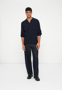 Marineblauwe ribgebreide pullover met een puntige kraag, gecombineerd met donkere, gestructureerde wijd beenbroeken en zwarte schoenen. Eenvoudig en modern ontwerp.