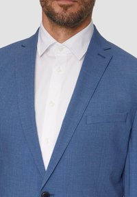 Hele sinine bleiser tekstuuriga kangast, kitsaste reveridega, rinnatasku ja nööpidega kinnitamine, koos valge kraega särgiga.