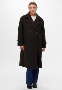 Sieviete, tērpusies garā melnā divrindu mētelī virs gaišā džemperī, zilu džinsu un melnām zābakiem, stāvot pretī baltai fona.