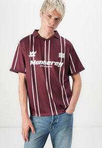 Bordeaux kortermet skjorte med hvite vertikale striper, krage og "Monterrey"-logo. Kombinert med blå denimjeans, poserer modellen mot en ensfarget bakgrunn.