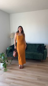 Vestido maxi amarelo mostarda, justo, com decote quadrado. Bolsa castanha e sandálias plataforma castanhas. Sofá de veludo verde, piso de madeira, planta nas proximidades.