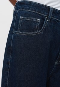 Los vaqueros de mezclilla azul oscuro tienen un corte clásico, costuras blancas en contraste y un bolsillo trasero con un borde limpio y recto.