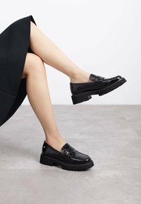 Mocasines de charol negros con una suela gruesa, que presentan una textura suave y sutiles detalles de costura, colocados sobre una superficie gris neutro.