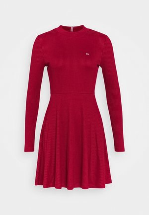 Robe rouge à manches longues et à côtes, avec corsage ajusté, jupe évasée et petit logo sur la poitrine gauche, présentée sur un fond blanc.