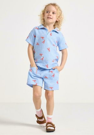 Blond child wearing light blue shorts and shirt set with red lobster prints, pink-striped socks, and brown sandals, walking with hands in pockets.