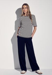 Camisa de rayas de manga corta en blanco y negro, combinada con pantalones holgados en azul marino y zapatillas blancas. El modelo está de pie con las manos en los bolsillos.