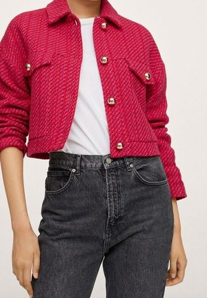 Felroze gewatteerde jas met twee borstzakken en gouden knopen, gedragen over een wit shirt en gecombineerd met zwarte high-waisted jeans.