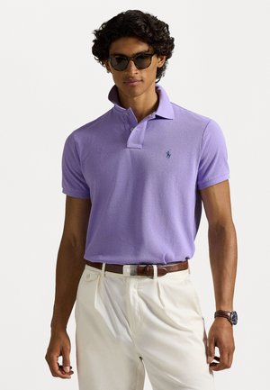 Fioletowa koszulka polo z teksturowanej tkaniny i kołnierzykiem zapinanym na dwa guziki, w połączeniu z kremowymi spodniami i brązowym paskiem. Widoczny detal z logo na piersi.