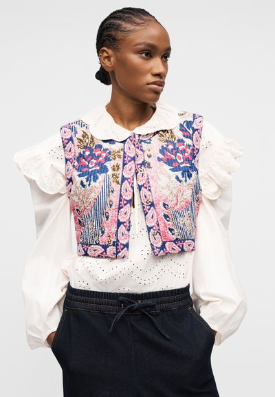 Femme aux cheveux tressés portant un chemisier blanc en broderie anglaise avec des manches volantées et un gilet sans manches orné de broderies florales roses et bleues.