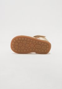 Sandale marron clair avec semelle en caoutchouc texturé présentant des motifs ronds. Fermeture à bande velcro et accent brillant sur le matériau supérieur.