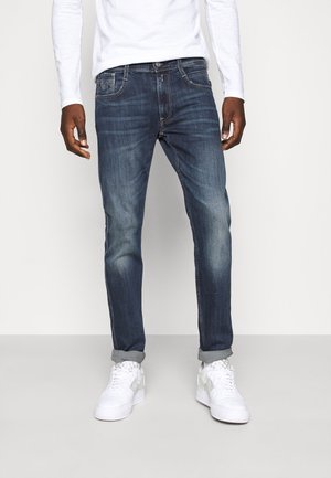 Homme portant un jean bleu foncé retroussé, une chemise blanche à manches longues et des baskets blanches, debout devant un fond uni.
