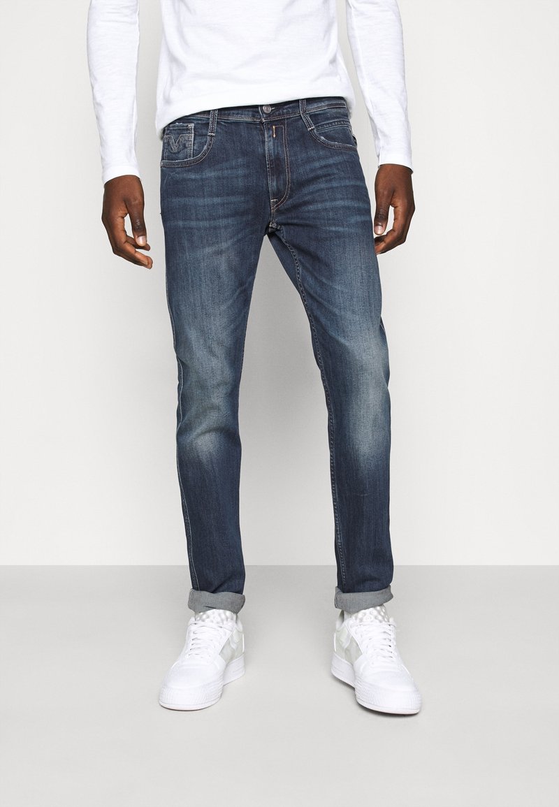 Homme portant un jean bleu foncé retroussé, une chemise blanche à manches longues et des baskets blanches, debout devant un fond uni.