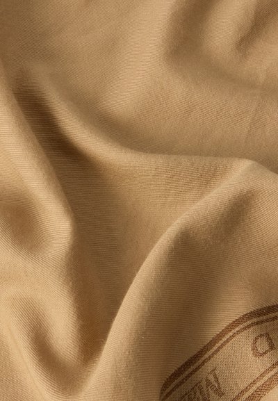 Tissu beige avec des plis doux et une texture fine côtelée, montrant une partie d'un motif ou texte imprimé brun sur le bord inférieur droit.