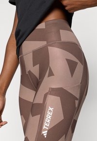 Leggings à motif géométrique marron en tissu extensible, avec une poche latérale et le logo "TERREX" en blanc sur la partie inférieure de la jambe.