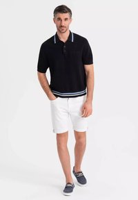 Polo negro con un cuello y dobladillo a rayas en azul claro, combinado con pantalones cortos blancos remangados y zapatos de barco azul marino. Presenta un bolsillo en el pecho.