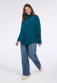 Poncho oversize in maglia turchese con polsini a costine, abbinato a jeans a zampa di colore azzurro chiaro. Scarpe da ginnastica con suola texturizzata completano il look.