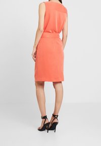 Robe sans manches corail avec une taille cintrée, une texture lisse et une coupe droite, associée à des sandales noires à talons hauts et brides.