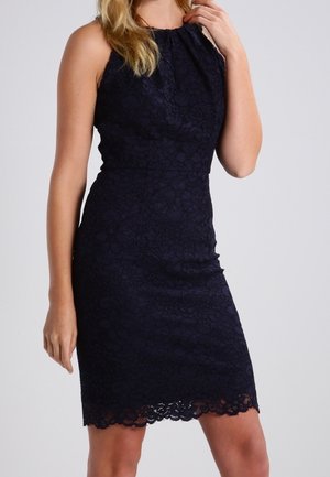 Femme portant une robe noire en dentelle sans manches avec un col haut et un ourlet festonné, posant devant un fond uni.