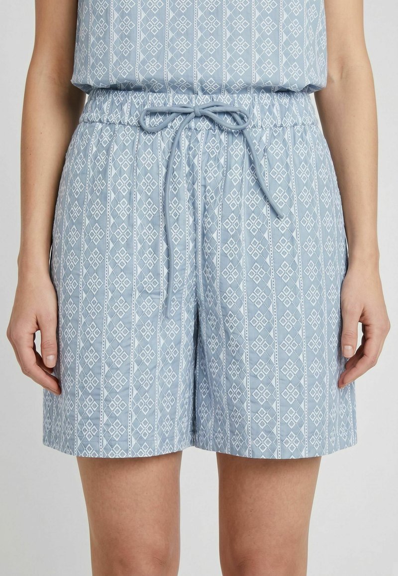 Shorts bleu clair avec un motif géométrique blanc en forme de losange et une taille à cordon, porté par une personne avec les bras détendus le long du corps.