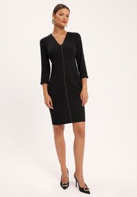 Czarna sukienka do kolan z dekoltem w kształcie litery V i rękawami o długości trzech czwarte. Posiada dekoracyjne złote guziki na przodzie.